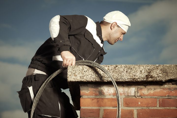 Chimney sweep technician in work uniform cleaning brick style chimney on building roof